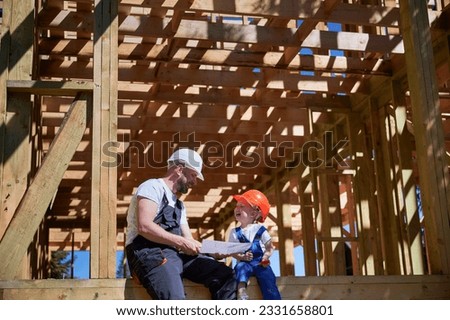 Father with toddler son building wooden frame house. Boys having fun on the edge of balcony, examining the construction plan, wearing helmets and overalls on sunny day. Carpentry and family concept. Royalty-Free Stock Photo #2331658801