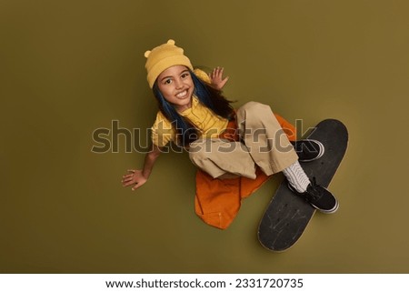 Top view of smiling and stylish preteen girl with colored hair wearing hat and urban outfit while looking at camera and sitting near skateboard on khaki background, girl with cool street style look Royalty-Free Stock Photo #2331720735