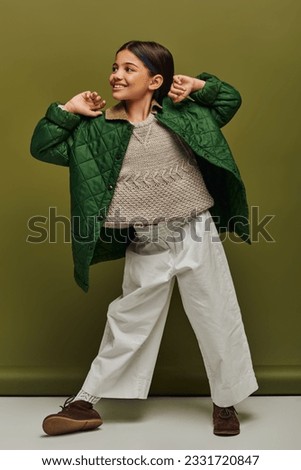 Full length of smiling and trendy preteen kid in autumn outfit and knitted sweater looking away while posing and standing on green background, modern child fashion for preteens concept Royalty-Free Stock Photo #2331720847