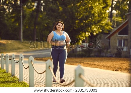 Happy overweight woman jogging in park. Smiling motivated beautiful fat large plump stout lady in sports top bra and yoga pants running on paved park path on sunny morning or evening. Fitness concept Royalty-Free Stock Photo #2331738301