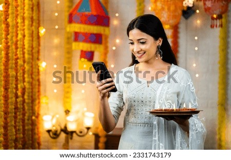 Happy young indian girl using mobile phone by holding diya lamp plate during festival celebration at home - concept of diwali offers, social media sharing and checking wishes or greetings. Royalty-Free Stock Photo #2331743179