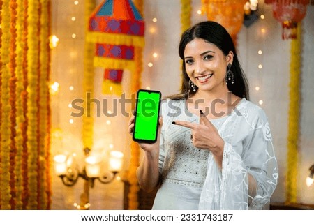 Happy young indian girl showing green screen mobile phone by pointing finger by looking camera - concept of festival offers, sales and app or application promotion. Royalty-Free Stock Photo #2331743187