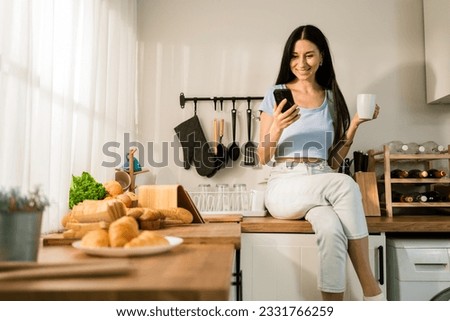 An attractive Caucasian woman using a smartphone while relaxing, drinking coffee and sitting on the kitchen counter at home in the morning. Happy woman shopping on mobile phone while in the kitchen.