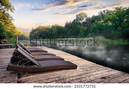 Wicker chaise longues near river at foggy sunrise