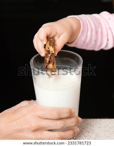 Caucasian girl dipping chocolate chip cookie into glass of milk.