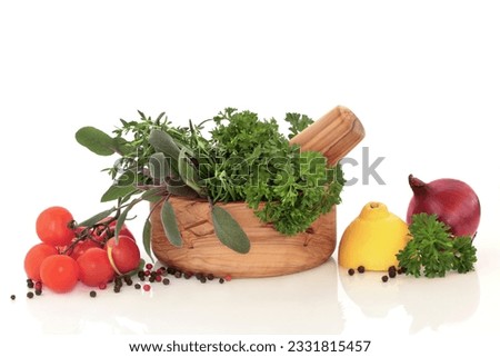 Herb leaf selection in an olive wood mortar and pestle, with lemons, red onion halves, tomatoes on the vine and peppercorns, over white background.