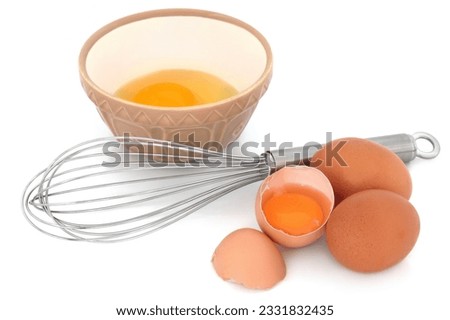 Metal whisk with speckled eggs and a raw egg in a baking mixing bowl, over white background.