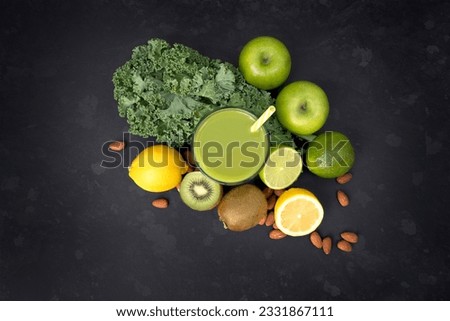 Healthy living concept with a glass of green smoothie surrounded by Kale, apples, almonds, limes and other fruit and veg on a dark background.