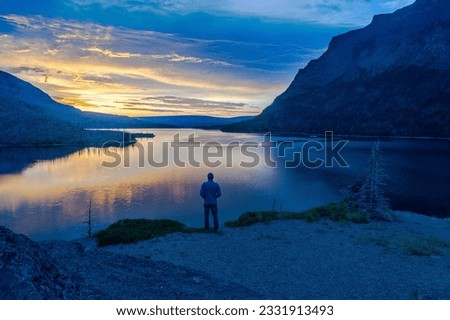 Man standing, looking away at a mountain lake and the sun rising behind a distance hills and clouds, Sun Point, St Mary Lake, Glacier National Park, Montana Royalty-Free Stock Photo #2331913493