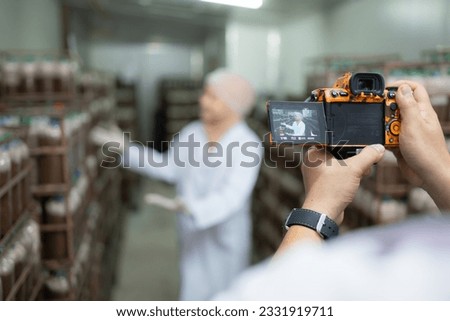 A photographer holding a digital camera on a production line is recording the work of a mushroom house worker. To be used for public relations and further marketing