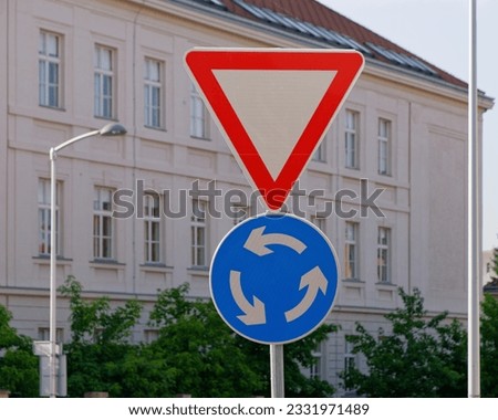 Close-up photo of a yield and roundabout traffic sign attached to a metal pole with the building in the background