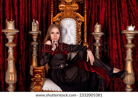 Cute lady in historical image sitting on antique throne fortress, looking at camera. Medieval queen in an old style posing on golden throne in castle room. Concept of theatre perform. Copy text space