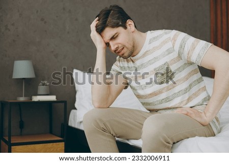 Side view young disappointed frustrated sad depressed man wears casual clothes t-shirt pajama sitting on bed prop up head spend time in bedroom home in own room hotel wake up dream be in bad mood day Royalty-Free Stock Photo #2332006911