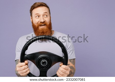 Young smiling fun redhead bearded man wear violet t-shirt casual clothes hold steering wheel driving car look aside isolated on plain pastel light purple background studio portrait. Lifestyle concept