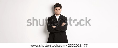 Portrait of angry handsome man in black suit, cross arms on chest and looking offended, frowning and pouting, being mad at someone, standing over white background. Royalty-Free Stock Photo #2332018477