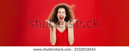 Woman releasing stress screaming out loud. Expressive and over-emotive attractive curly-haired woman in elegant red dress yelling with closed eyes and palms raised near face over studio background