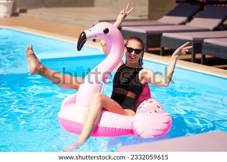 Hot slim woman have fun and wave hand on inflatable pink flamingo float mattress in bikini at swimming pool. Attractive fit girl in swimwear lies in sun on floaty. Pretty female on tropical vacation.