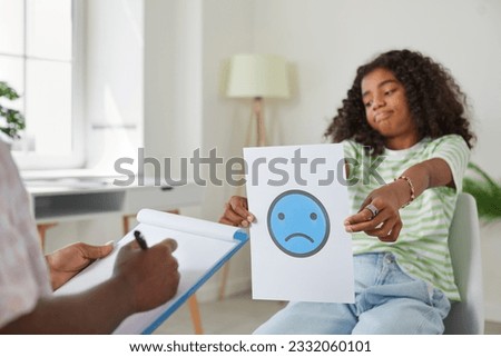 Sad, stressed African American school child girl sitting on chair and holding picture of unhappy emoji while therapist or school psychologist is writing on her clipboard. Children's psychology concept