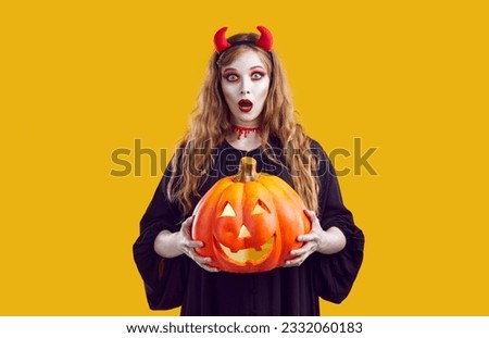 Surprised girl with Halloween makeup holding jack-o-lantern. Beautiful amazed teenager wearing red devil horns and black dress standing over isolated studio background with mouth wide open