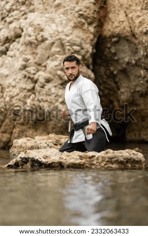 Martial arts master in defensive position between rocks in the sea with the water at knee height wearing white kimono, black trousers and a black belt on which is written the word "Bushido" in Japanes Royalty-Free Stock Photo #2332063433