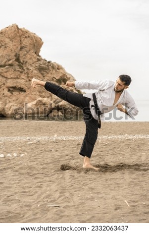 man practising martial arts in a kimono with a black belt with the word "Bushido" in Japanese as he performs a side kick on the sand on the beach. Royalty-Free Stock Photo #2332063437