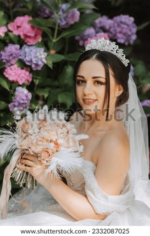 young beautiful bride in wedding dress with open shoulders and crown on her head sitting near hydrangea flowers, fashion photo taken under harsh sunlight