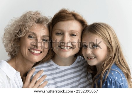 Cheerful cute little kid girl, pretty mom and grandma standing close, hugging with head touches, looking at camera with toothy smiles, promoting family dental care, posing at white wall