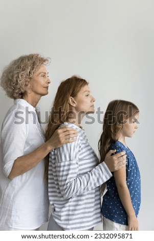Pretty young mom, grandma and cute preschool kid girl posing for picture at white wall background, standing behind each other, touching shoulders of daughters, looking forward. Vertical portrait