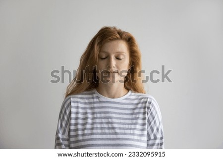 Calm red haired young woman standing at white wall background with closed eyes, sleepy face, taking deep breath, meditating. Peaceful tranquil beautiful model head shot portrait