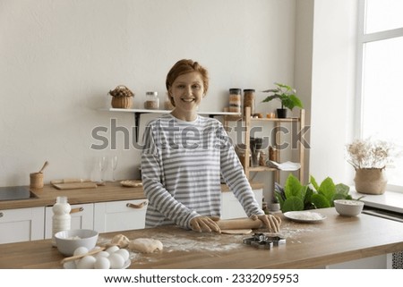 Happy pretty young baker woman rolling cooking in home kitchen, rolling dough on table with flour, eggs, milk, baking cookies, biscuits, dessert snacks, looking at camera, smiling