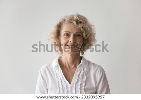 Positive beautiful retired woman standing at white wall background, posing for shooting, looking at camera, smiling. Pretty mature female model with blonde curly hair head shot portrait