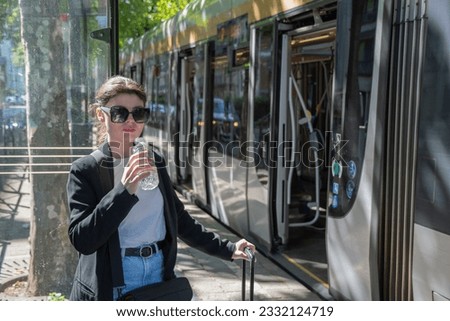 a business woman in sunglasses drinks water at a tram stop in the city, dressed