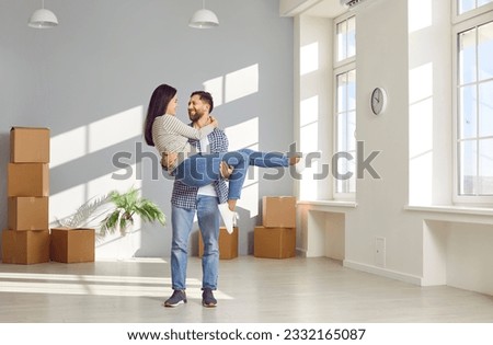 Young family couple having fun on moving day in new house or apartment. Happy husband carrying wife. Joyful man standing in empty unfurnished living room with boxes and holding his woman in his arms