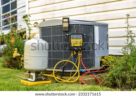 An old air conditioning unit that is being repaired by an HVAC service technician. The technician is using digital refrigerant gauges, and a digital scale.  Royalty-Free Stock Photo #2332171615