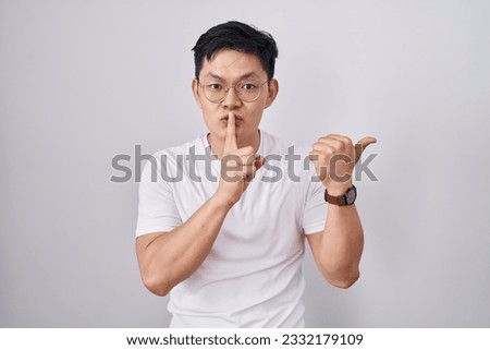 Young asian man standing over white background asking to be quiet with finger on lips pointing with hand to the side. silence and secret concept. 