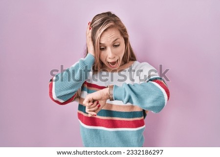 Young blonde woman standing over pink background looking at the watch time worried, afraid of getting late 