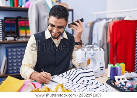 Young arab man tailor listening voice message by smartphone writing on notebook at clothing factory