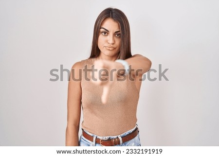 Young hispanic woman standing over white background looking unhappy and angry showing rejection and negative with thumbs down gesture. bad expression. 