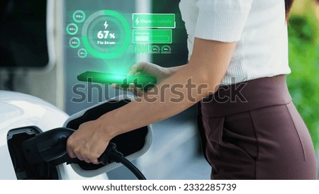 Modern lifestyle woman insert EV charger to her electric car at home charging station and check EV battery status hologram from smartphone. Futuristic energy sustainability and electric car. Peruse