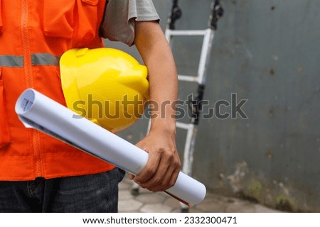 Front view of male construction worker holding blueprint and safety helmet outdoor of construction site with ladder in the background. Copy space and selective focus. Royalty-Free Stock Photo #2332300471