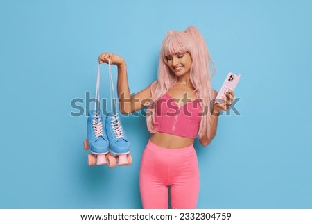 Girl in pink top and leggins with pink hair holding blue roller-skates and smiling while posing on blue background, fashion style concept, copy space