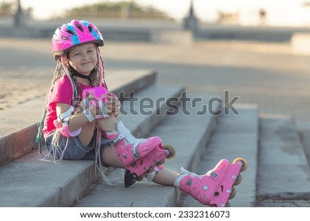 Happy little girl preschool in a bright pink summer t-shirt with colored pink-blue African braids in her hair on roller skates in a park. children's hobby. sports lifestyle