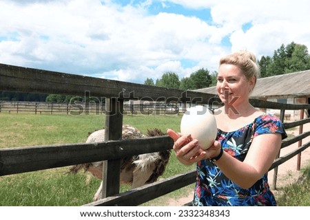 A woman holds an ostrich egg in her hands. Ostriches in a petting zoo. In a wooden cage.