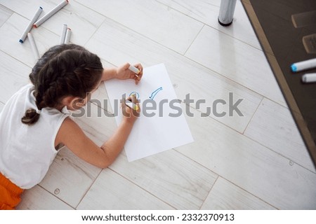 View from above of an adorable preschooler child girl in white t-shirt and bright orange shorts, is lying on the floor at home and drawing picture on a white paper sheet with watercolor felt-tip-pens