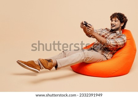 Full body young Indian man wear brown shirt casual clothes sit in bag chair hold in hand play pc game with joystick console isolated on plain pastel light beige background. People lifestyle concept