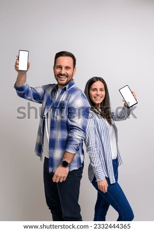 Smiling young couple standing back to back and showing blank screen of smart phones. Portrait of happy boyfriend and girlfriend promoting mobile apps against background