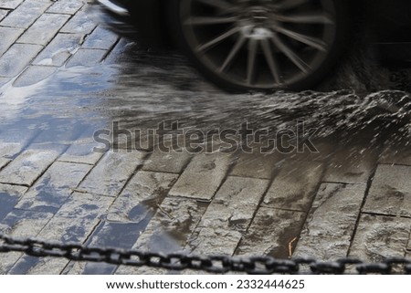 A close up photo of a car going through a puddle of water on a stone road with a chain in the foreground. 