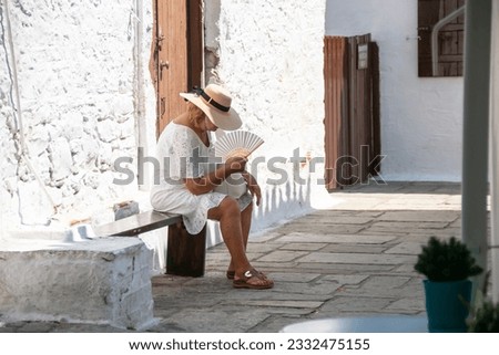 Heatwave image of a woman sitting down in the shade and cooling herself with a fan. Her head hangs low in the searing temperature. Selective focus on the woman. Royalty-Free Stock Photo #2332475155