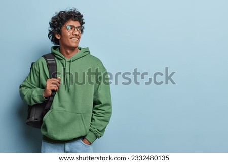Indoor waist up of young smiling happy Hindu man standing on left isolated on blue background wearing casual green hoodie and jeans with bag on left shoulder looking at blank space for your promotion Royalty-Free Stock Photo #2332480135