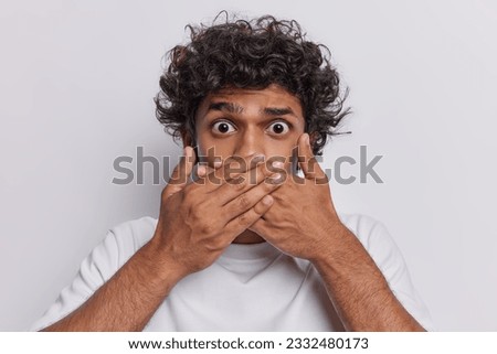 Keep silence. Stupefied curly haired Hindu man covering mouth with hands tries not to drop any word dressed casually isolated over white background. Speak no evil concept. Reaction to bad news